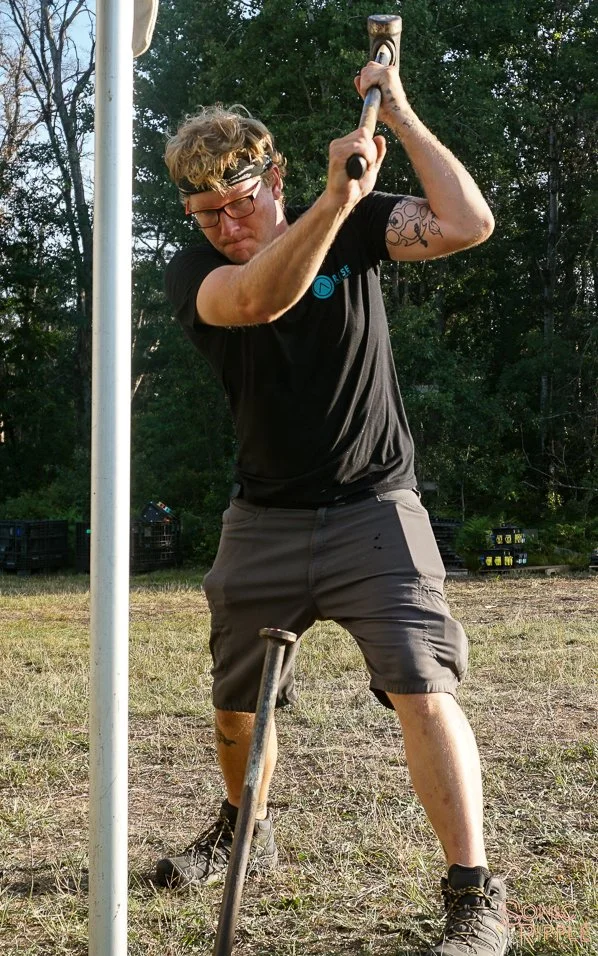 Man swinging a sledgehammer against a pole outdoors