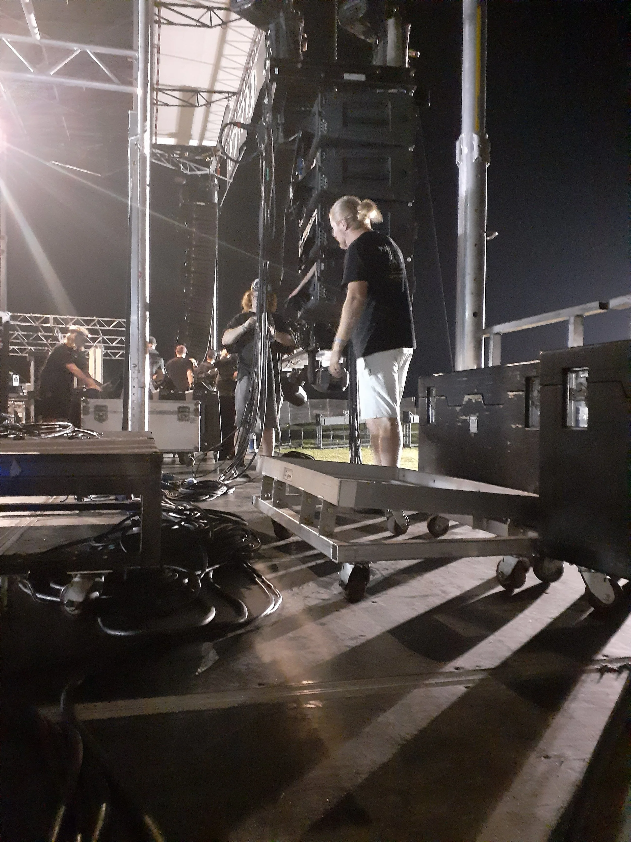 Crew setting up audio and lighting equipment at a nighttime outdoor concert stage