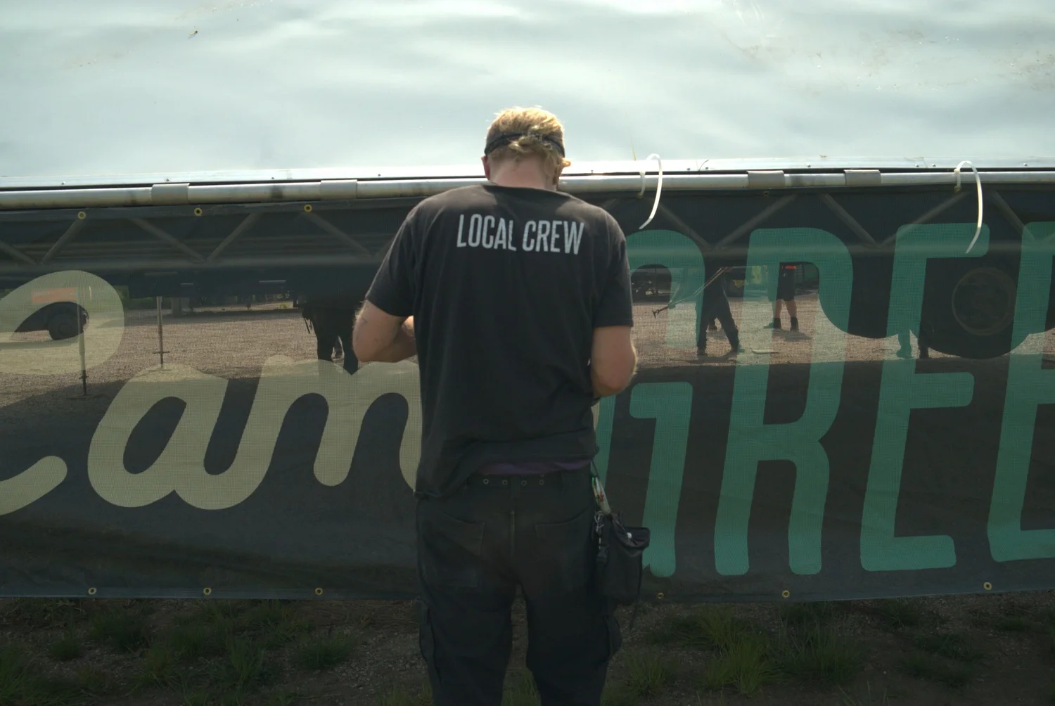 Person wearing a black T-shirt with LOCAL CREW written on the back, standing in front of a banner at an outdoor event